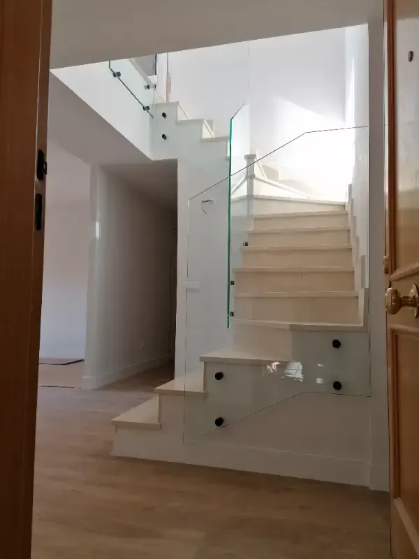 Un interior moderno con una escalera blanca con barandillas de cristal y un suelo de madera que se ve a través de una puerta.