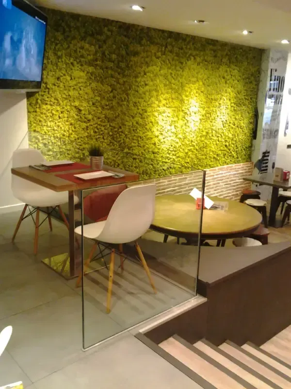 El interior de una cafetería moderna cuenta con una pared cubierta de musgo verde vibrante, mesas de madera y sillas blancas