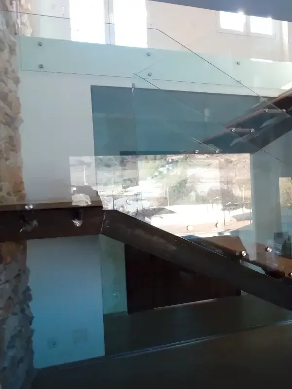 Vista de cerca de una barandilla de escalera moderna de cristal con pasamanos de metal, montada contra una pared de piedra.