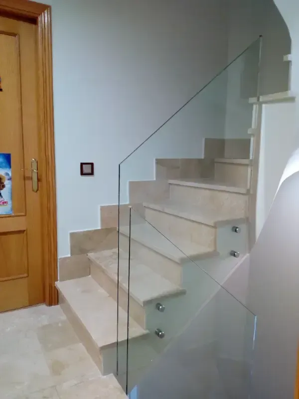 Escalera de piedra de color claro con barandilla de cristal sin marco, fijada con separadores metálicos en un pasillo.