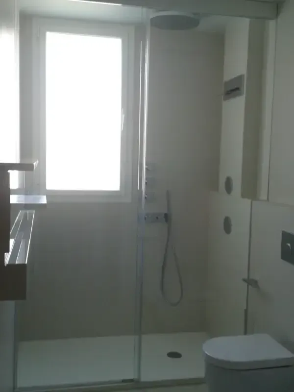 Un baño minimalista en tonos claros, con una cabina de ducha de cristal y un inodoro blanco moderno.