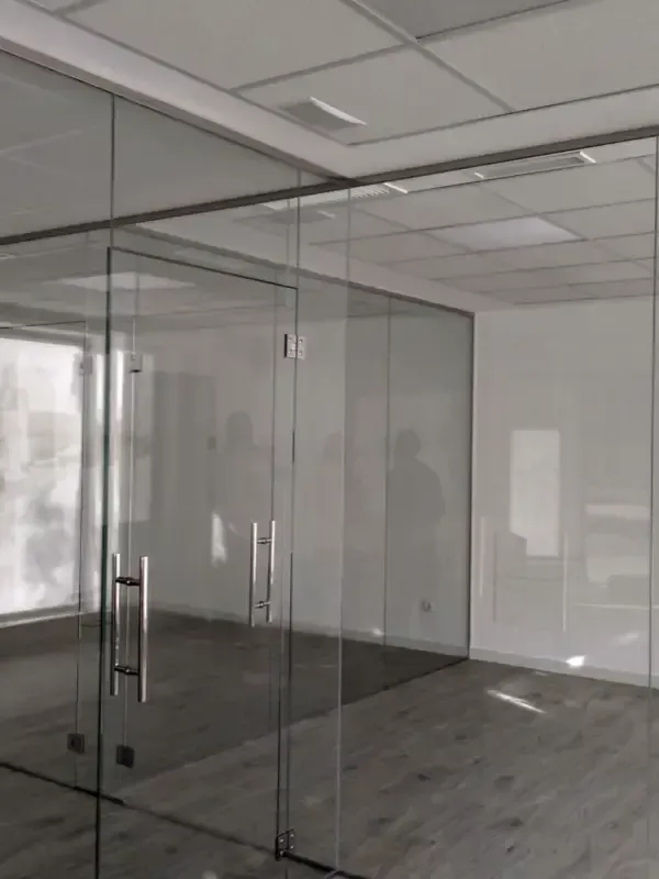 Mampara de oficina con paredes de cristal, puerta batiente de cristal, tiradores metálicos y suelo de imitación madera gris
