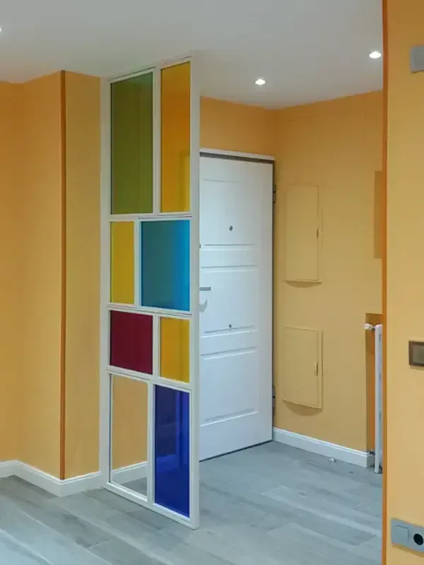 Un biombo con marco blanco y paneles de vidrio multicolor en verde, amarillo, azul y rojo, situado cerca de la entrada.