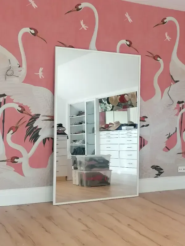 Un espejo de pie se apoya contra una pared con papel pintado rosa con estampado de grullas