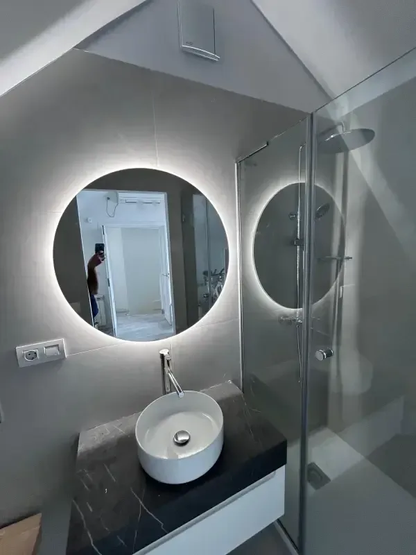 Baño moderno con espejo circular retroiluminado por LED, lavabo blanco sobre encimera de mármol oscuro y ducha de cristal.