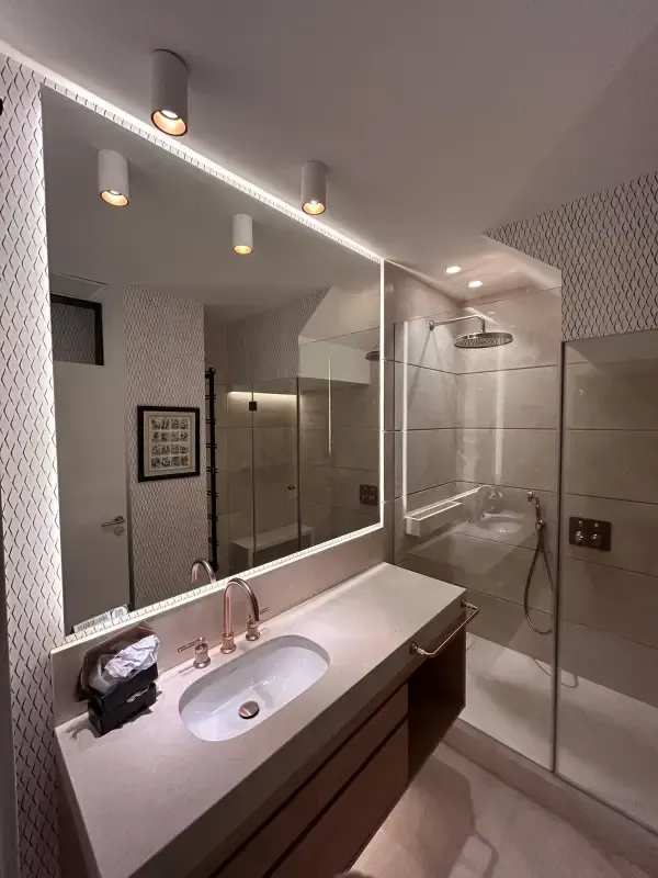 Un baño moderno con un tocador blanco con un solo lavabo, un gran espejo retroiluminado y mampara de cristal.