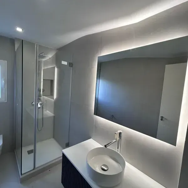Un baño moderno con ducha acristalada, lavabo blanco sobre encimera y un gran espejo rectangular retroiluminado.