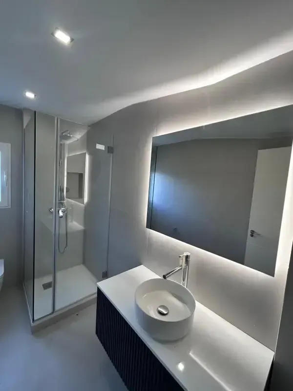 Baño moderno con lavabo blanco sobre encimera oscura, espejo de pared retroiluminado y ducha con mampara de cristal.