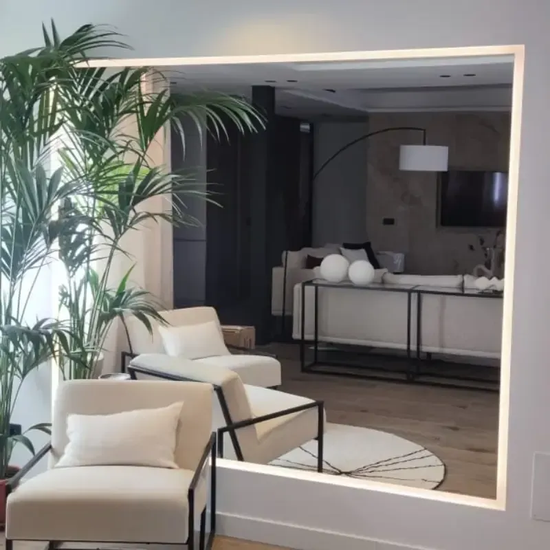 Un salón moderno reflejado en un gran espejo cuadrado retroiluminado, con dos sillones de color beige y una palmera