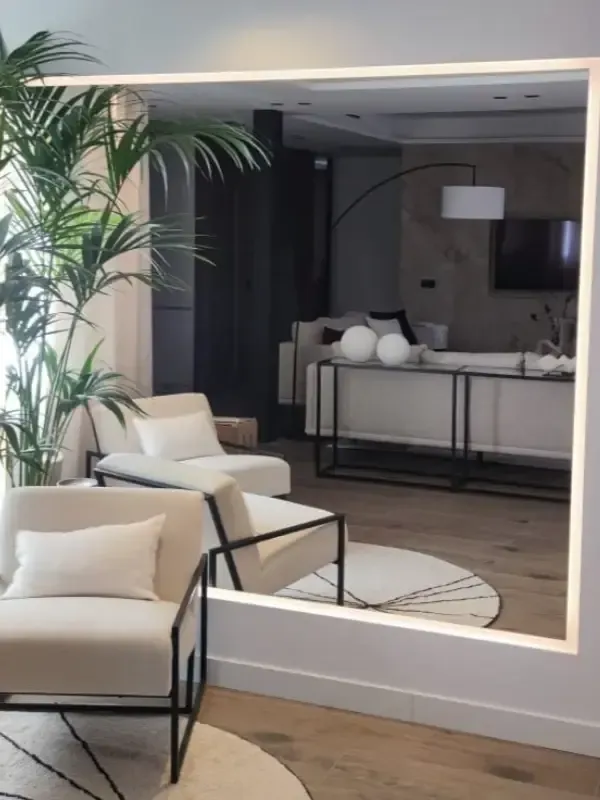 Un salón moderno reflejado en un gran espejo de pared retroiluminado, con sillones de color beige