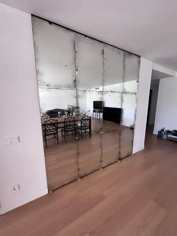 Una pared cubierta de grandes espejos antiguos rectangulares, que reflejan un comedor con sillas y un suelo de madera.