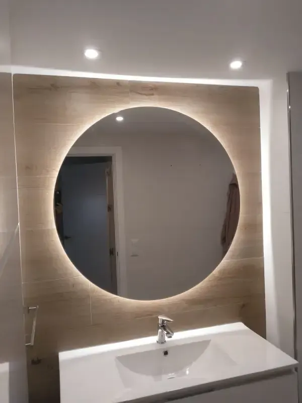Un moderno mueble de baño con un gran espejo circular retroiluminado, montado sobre una pared revestida con azulejos