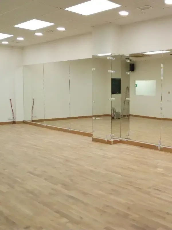 Un estudio vacío de fitness o danza con suelo de madera clara, paredes blancas y un gran espejo montado en la pared.