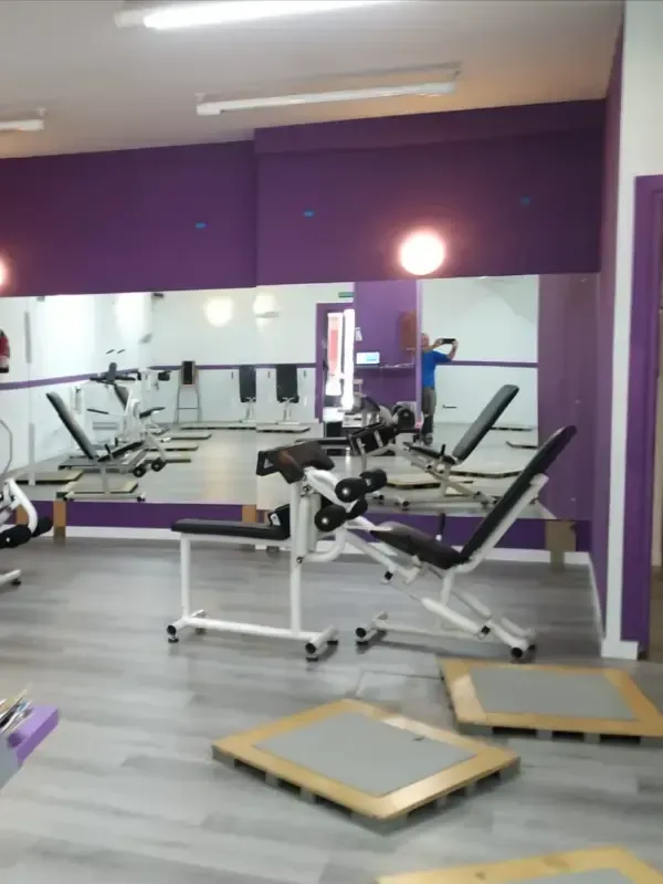 Interior de un gimnasio con paredes moradas, espejos, máquinas de ejercicio blancas y colchonetas en el suelo.