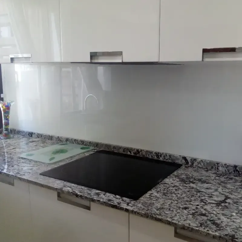 Una encimera de cocina con armarios blancos, una placa de inducción negra y un protector contra salpicaduras de vidrio.
