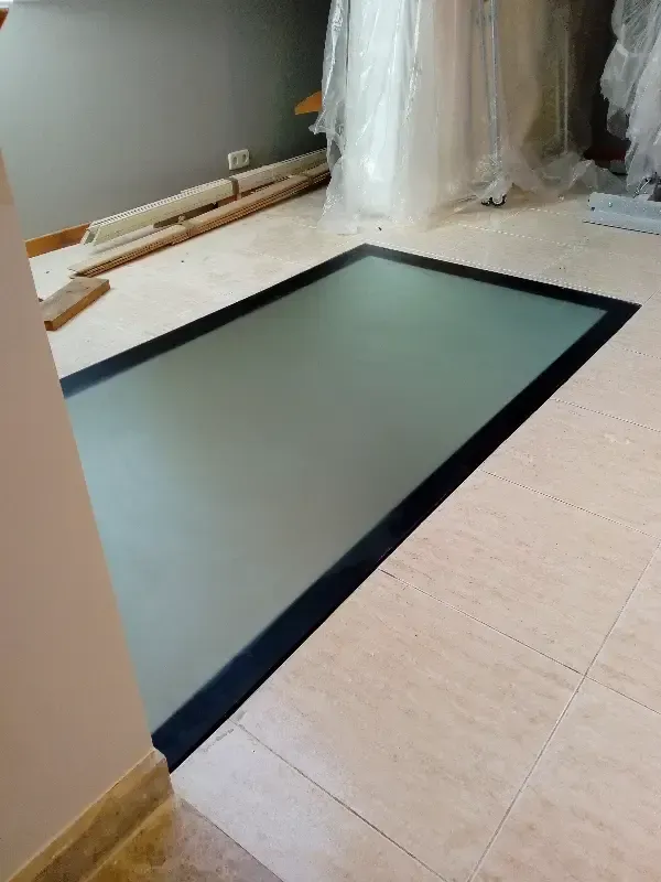 Un panel rectangular de vidrio esmerilado en el suelo, enmarcado en metal negro y rodeado de baldosas de color claro.