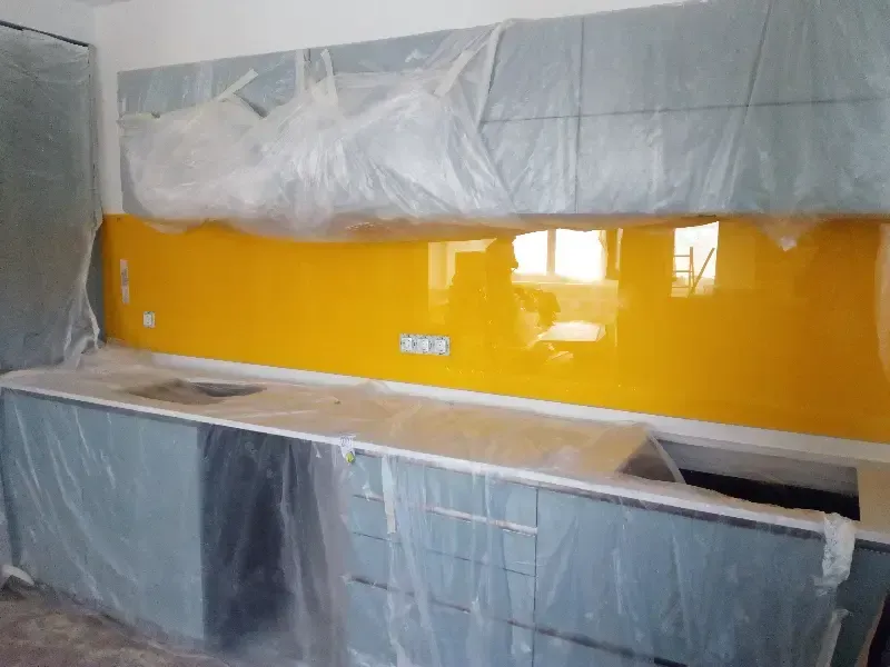 Una cocina en proceso de renovación con armarios grises, un protector contra salpicaduras de vidrio amarillo brillante