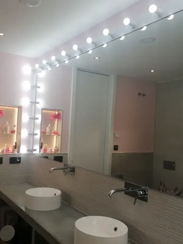 Tocador de baño moderno con dos lavabos blancos, encimera gris y un gran espejo enmarcado por luces brillantes.