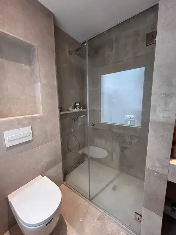 Un baño moderno en tonos neutros que cuenta con un inodoro blanco suspendido junto a una ducha con mampara de cristal