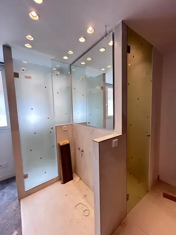 Un baño moderno en construcción con mamparas de vidrio esmerilado, un espejo grande e iluminación empotrada en el techo.