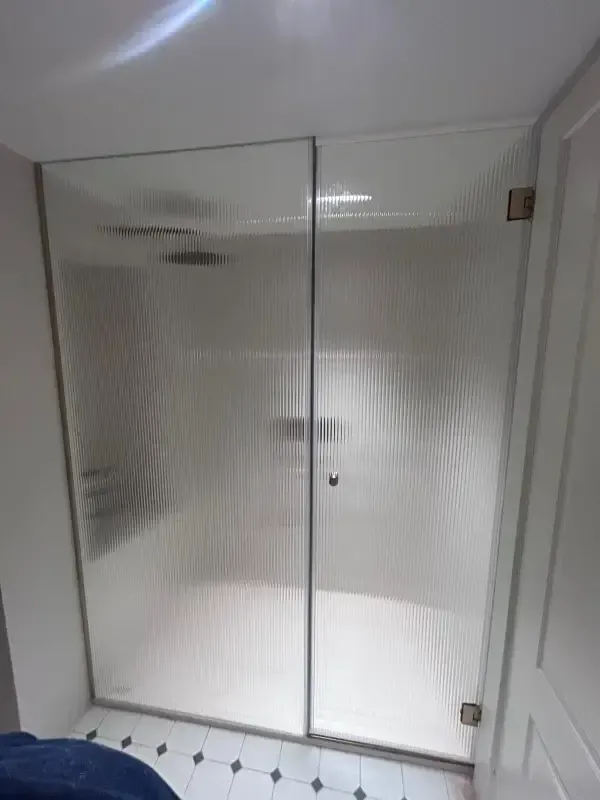 Una cabina de ducha de cristal con textura estriada vertical, grifería cromada y suelo blanco, instalada en un baño