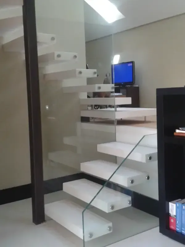 Escalera flotante con escalones blancos y barandilla de cristal en el interior de una habitación moderna de tonos neutros.