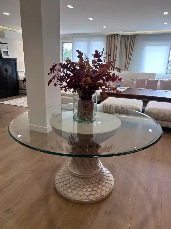 Una mesa de comedor redonda de cristal con una base de pedestal texturizada, que sostiene un jarrón con flores secas