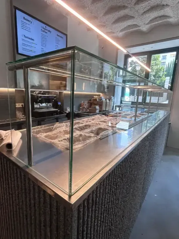 Un moderno mostrador de cafetería con vitrina de cristal, base gris texturizada y una pizarra montada en la pared trasera.
