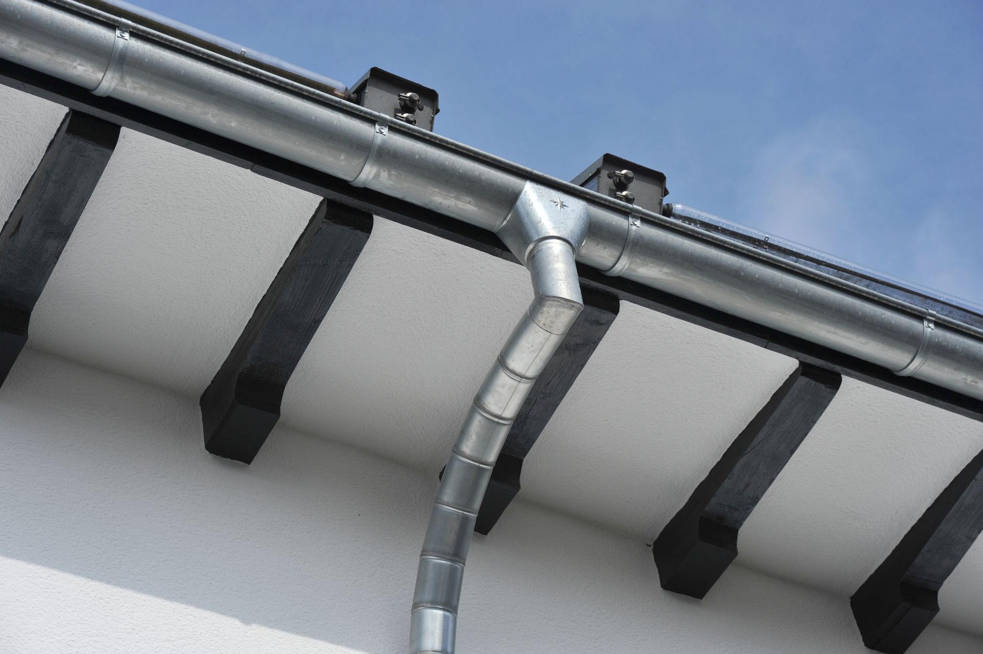 Gros plan sur un système de gouttière métallique avec descente pluviale fixée à un bâtiment blanc.