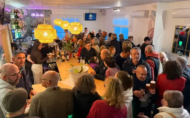 Recepción en un espacio interior abarrotado, con invitados charlando alrededor de largas mesas bajo cálidas luces colgantes.