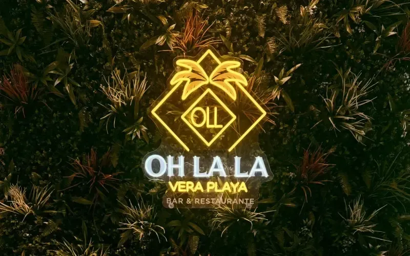 OH LA LA Logotipo de Vera Playa en dorado brillante sobre un fondo oscuro con hojas