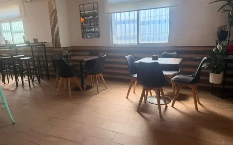 Interior vacío de una cafetería con mesas, sillas, grandes ventanales y una planta en maceta.