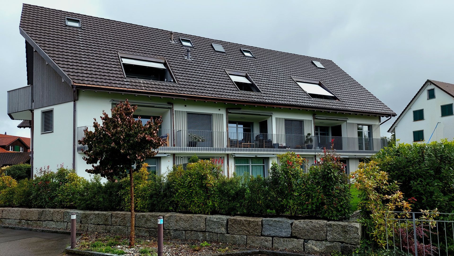 Ein großes weißes Haus mit braunem Dach ist von Bäumen und Büschen umgeben.