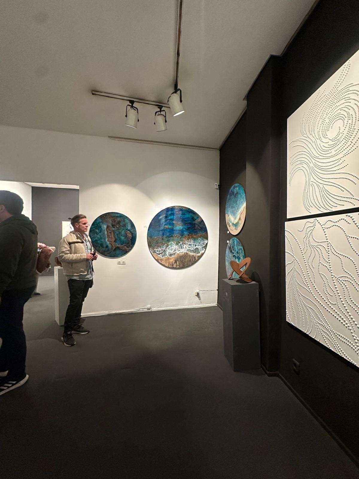 Galería de arte con pinturas circulares azules sobre paredes blancas y dos personas observando la exposición.