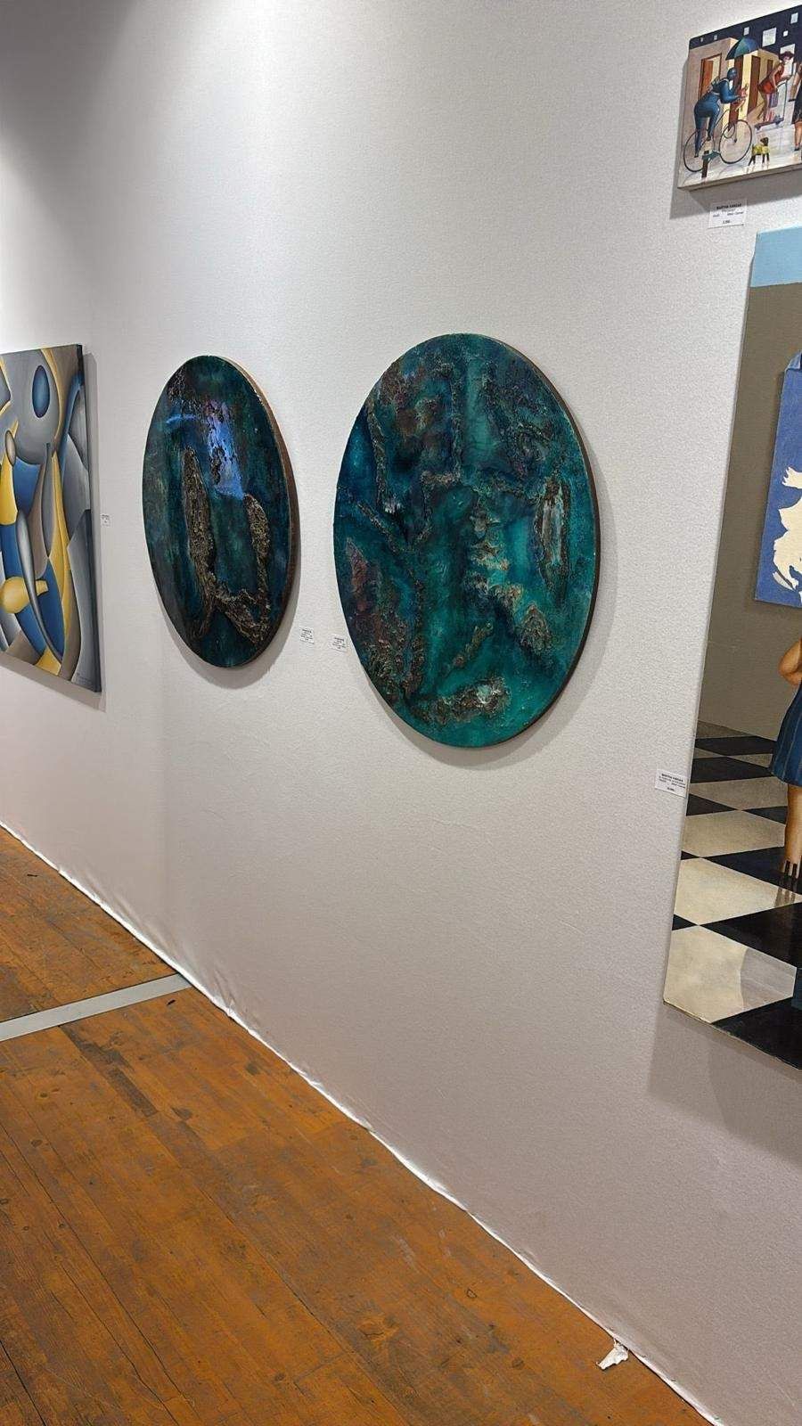 Pared de una galería de arte con tres pinturas ovaladas abstractas de color verde azulado y negro expuestas en fila.