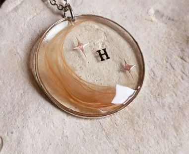 Collar con colgante transparente con un remolino dorado, una "H" negra y dos pequeñas estrellas rosas sobre un fondo claro.