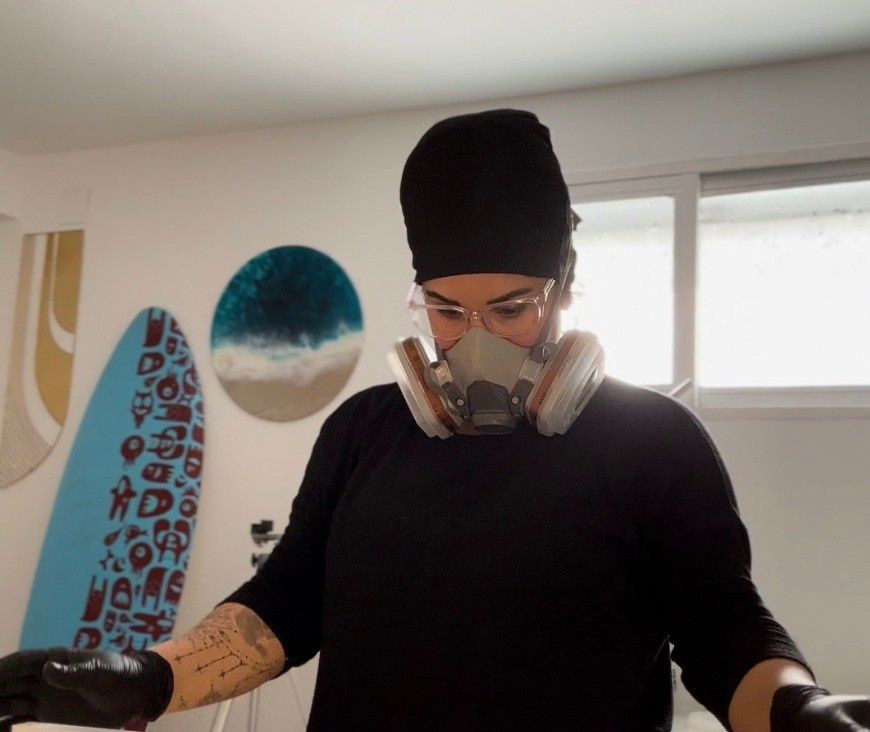 Persona con camisa negra y mascarilla de pie en el interior de un edificio, cerca de tablas de surf y una ventana.