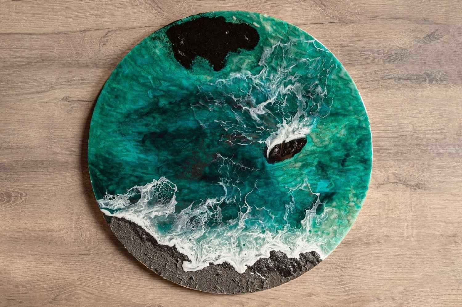 Obra de arte redonda de resina con forma de ola marina en color turquesa y blanco sobre un fondo de madera.