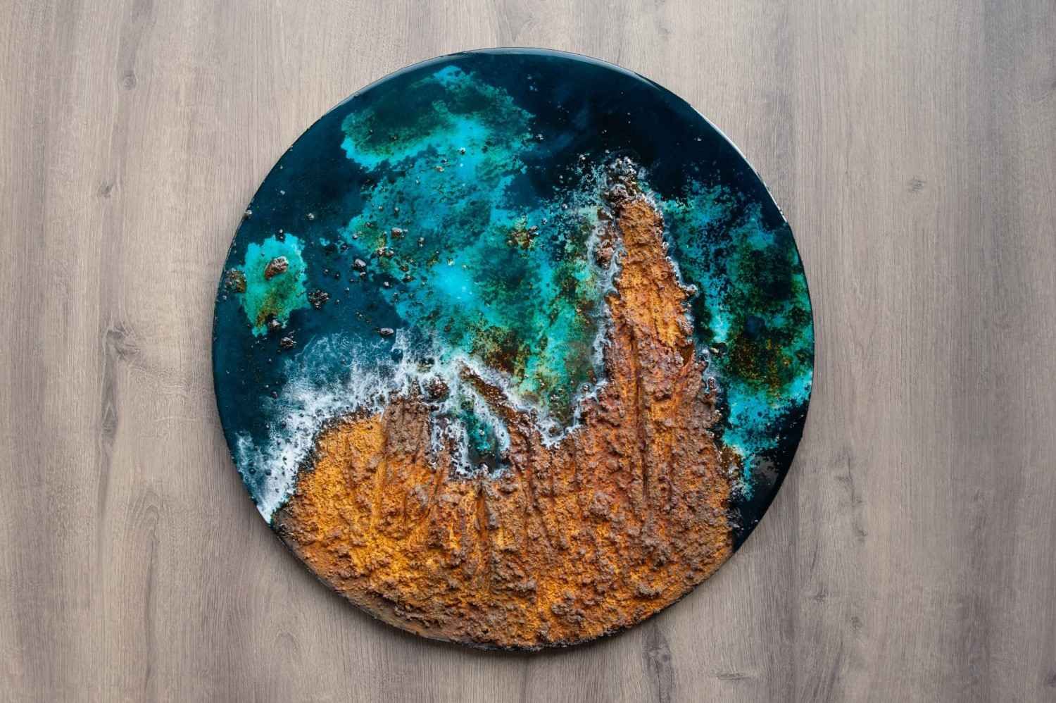 Obra de arte circular abstracta con texturas en tonos verde azulado, negro, blanco y naranja sobre una superficie de madera.