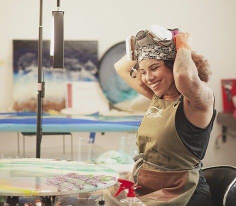 Artista sonriendo mientras se ajusta una peluca rizada junto a una mesa cubierta de pintura en un estudio.