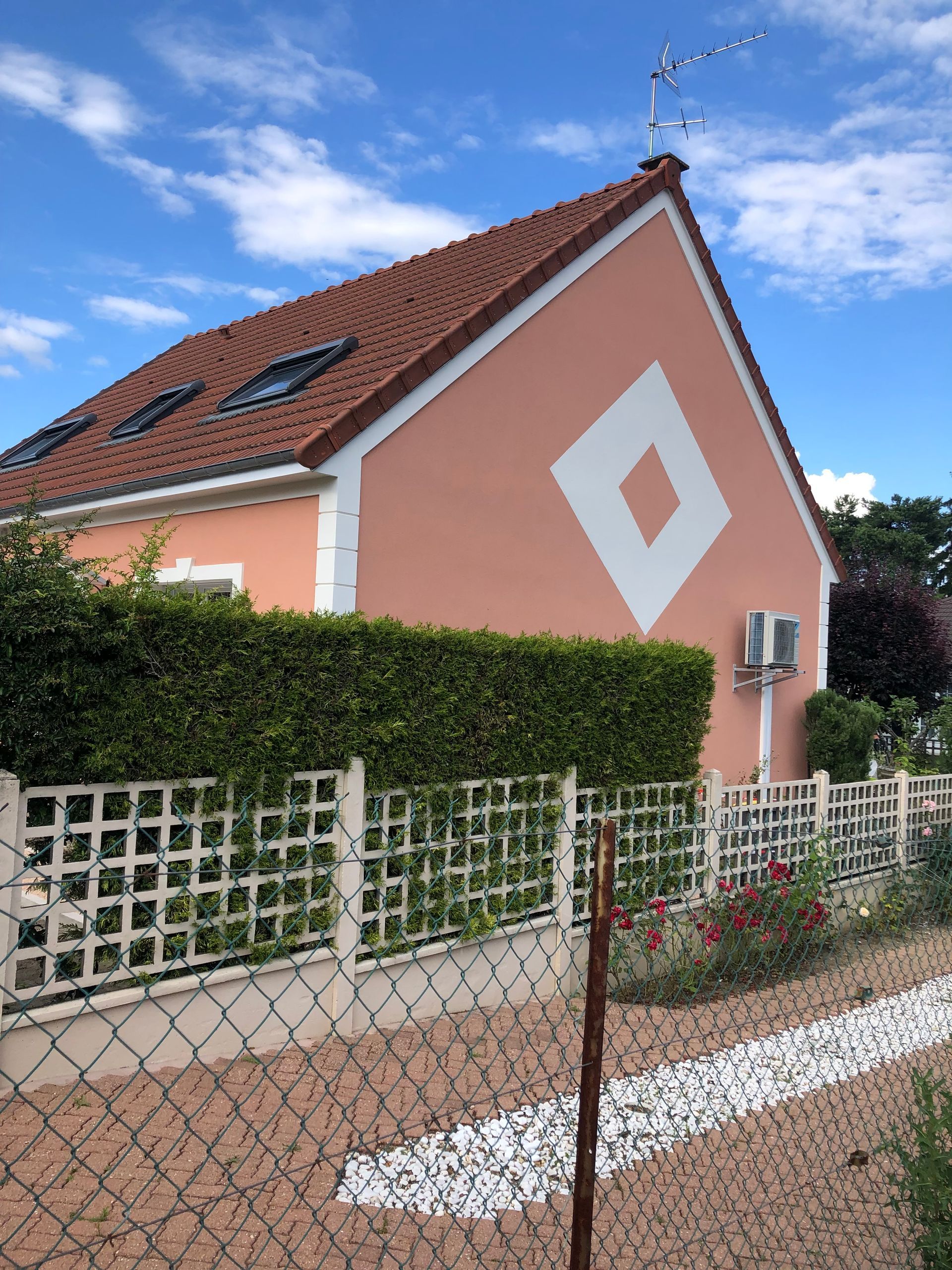 Maison rose au toit de tuiles rouges, avec des détails muraux en losanges blancs, une haie et une clôture grillagée sous un ciel bleu