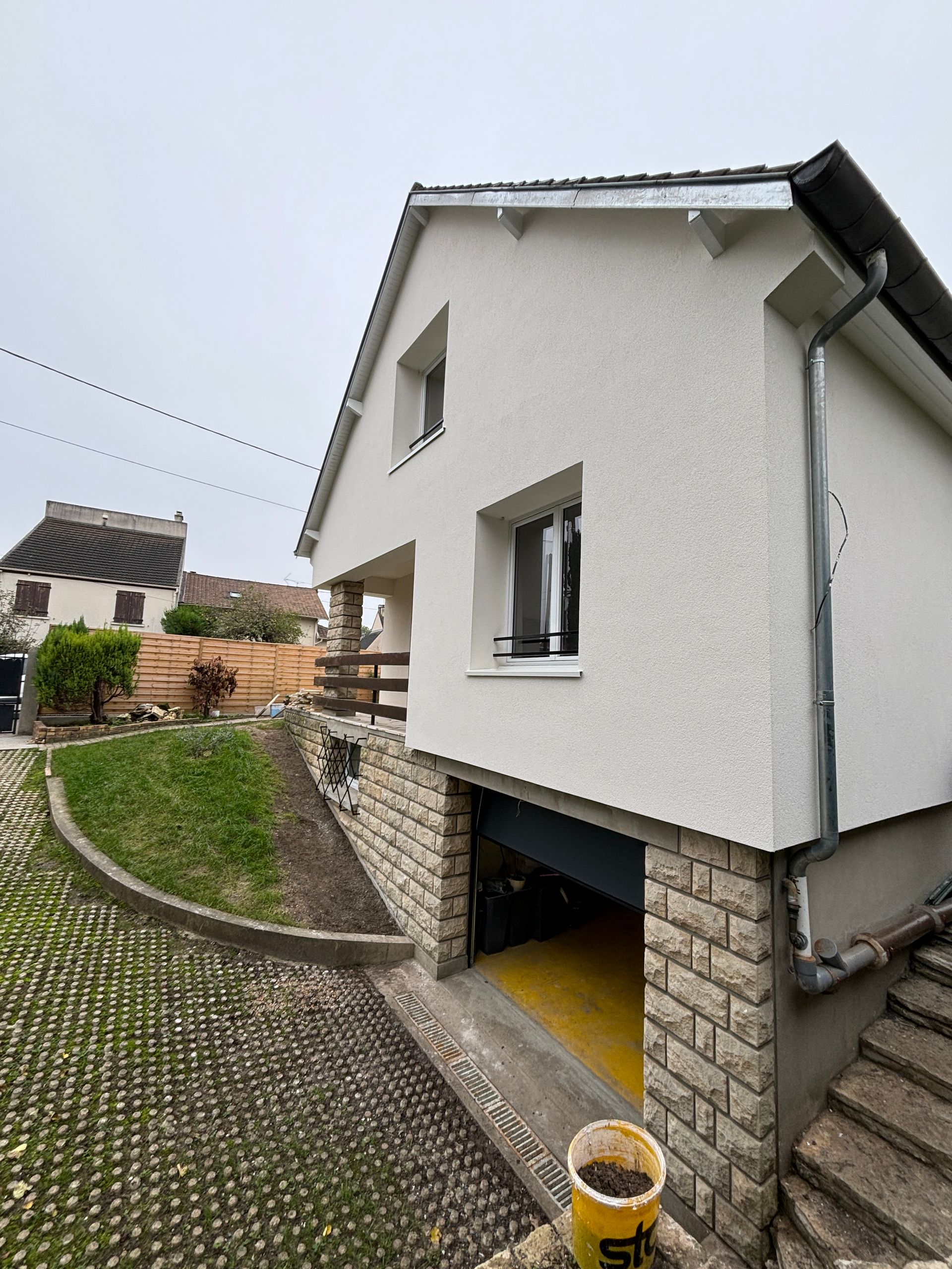 Maison blanche en stuc avec des fondations en pierre, située au bord d’une allée pavée en pente, à côté d’un chemin herbeux