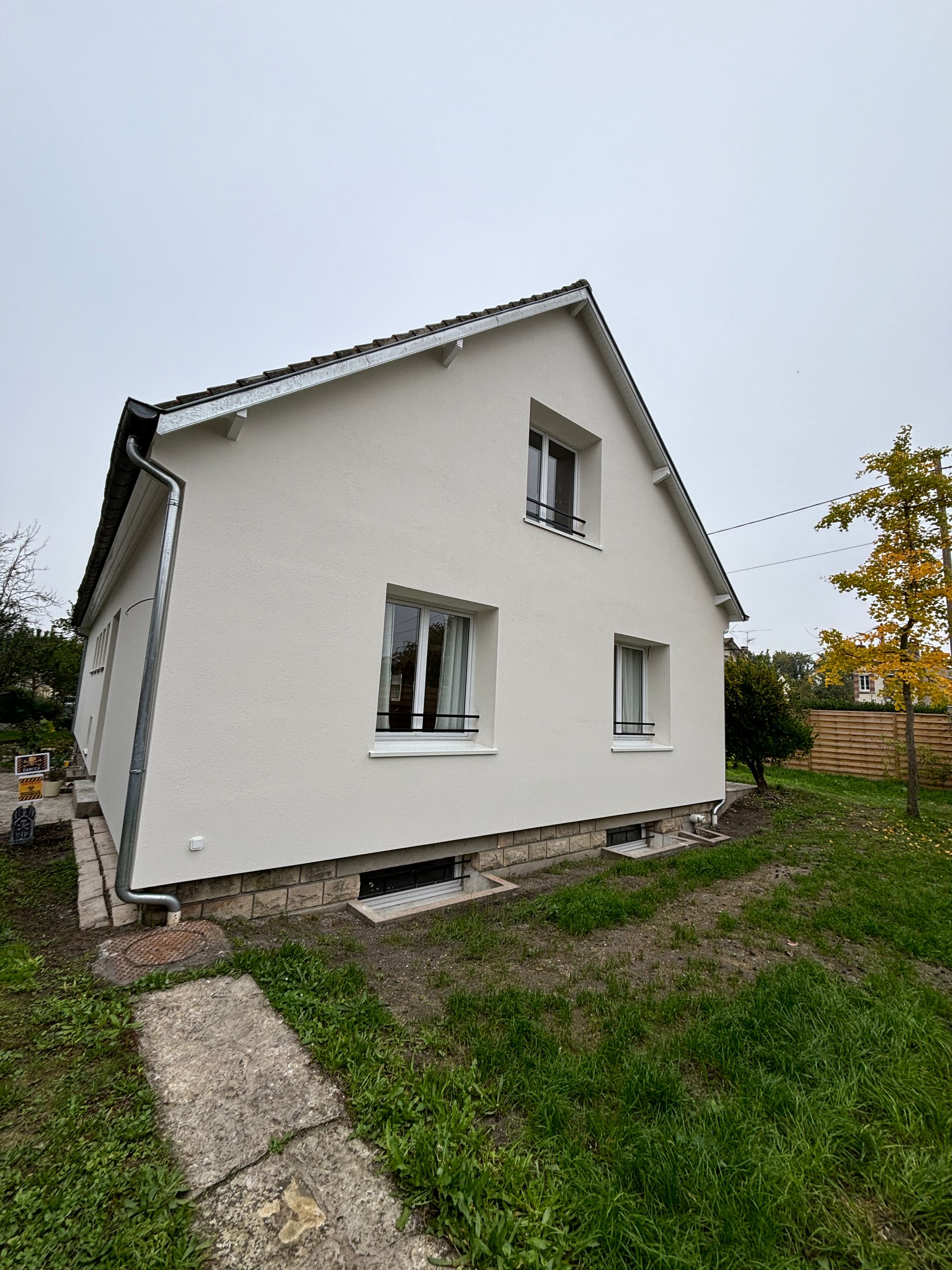 Maison blanche à deux étages, au toit pentu, avec des fenêtres et un jardin herbeux sous un ciel couvert.