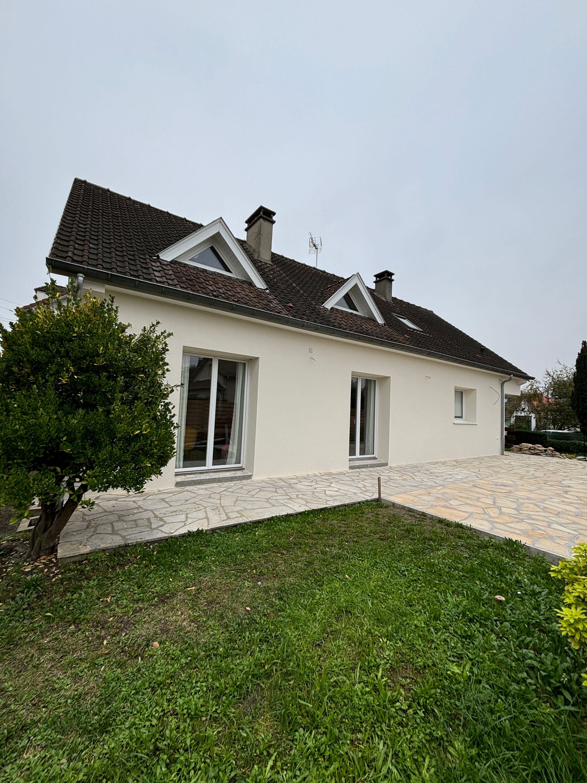 Maison blanche au toit sombre, avec de petites lucarnes, une pelouse verte et une allée en gravier, sous un ciel couvert