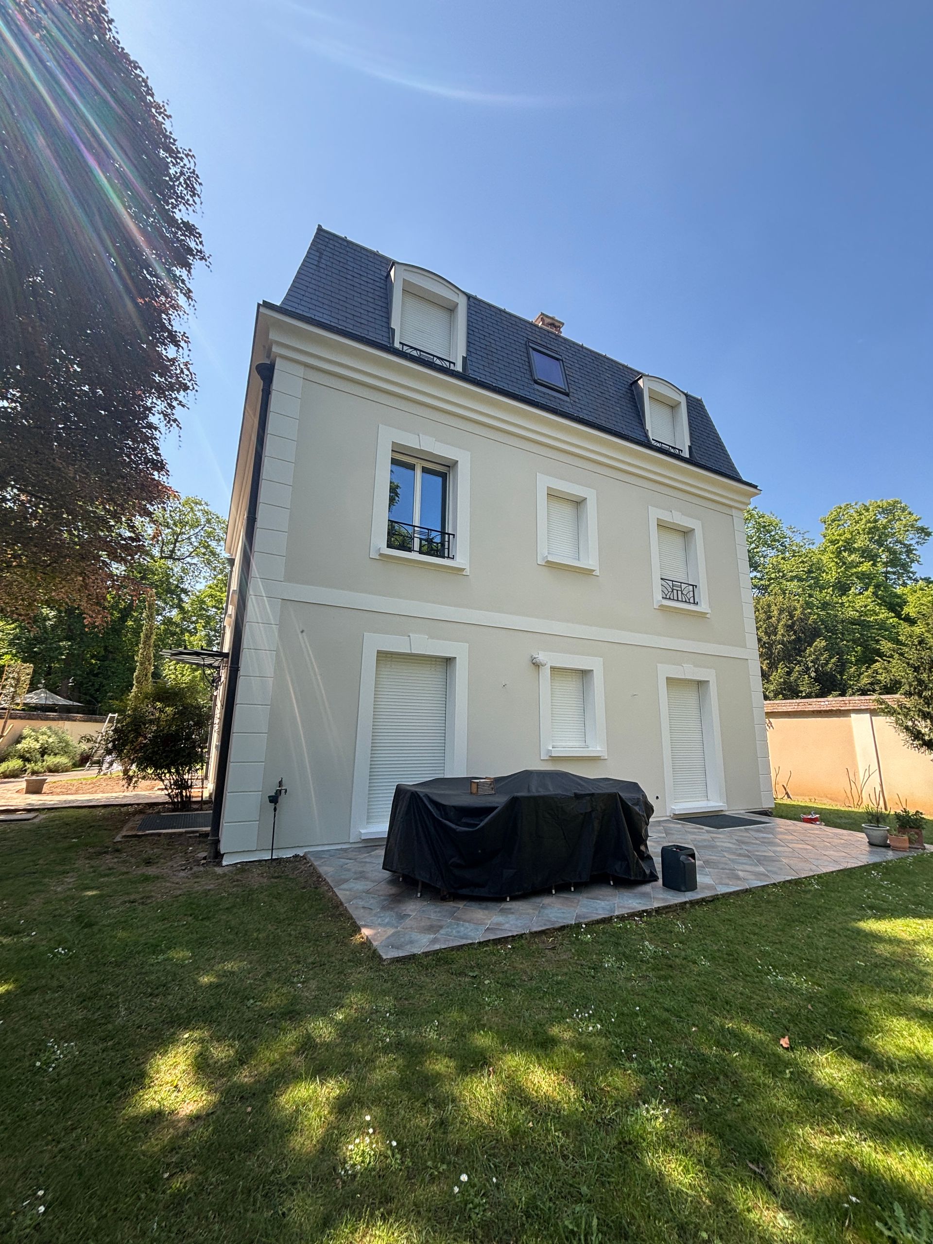 Maison blanche de trois étages, avec un toit sombre et des fenêtres à volets, vue depuis un jardin ensoleillé