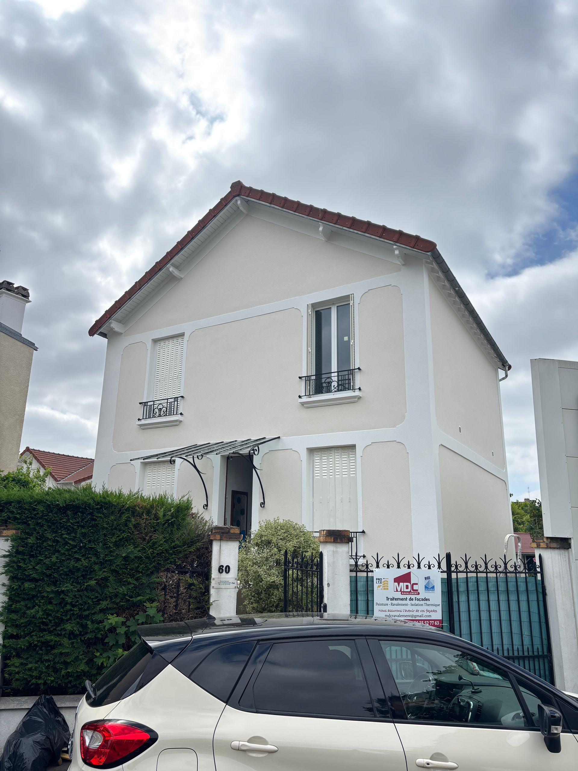Maison crème à deux étages avec un toit rouge, derrière une voiture noire garée sous un ciel nuageux