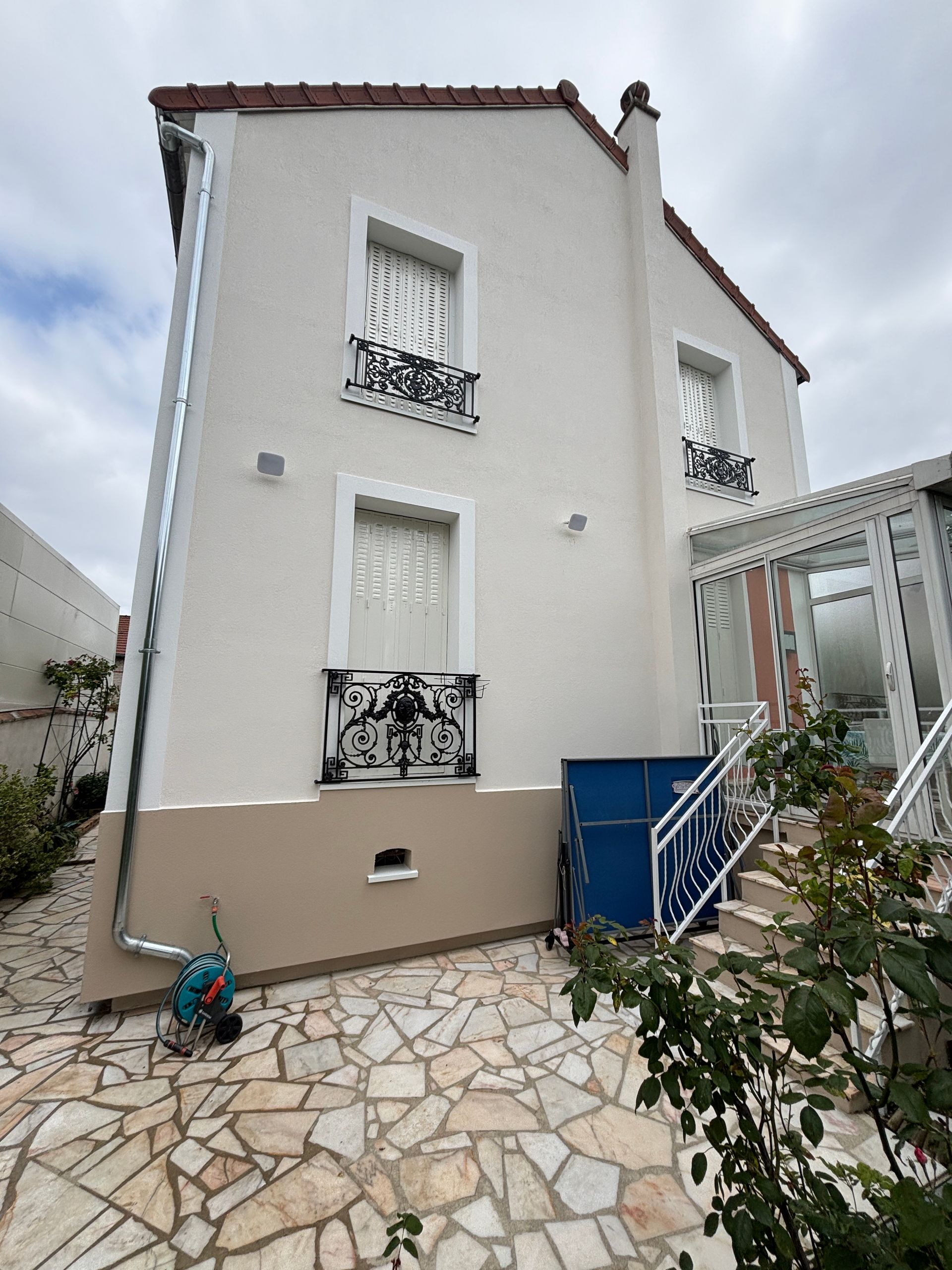 Maison blanche à deux étages avec des fenêtres à volets, un escalier bleu et une cour pavée de pierres