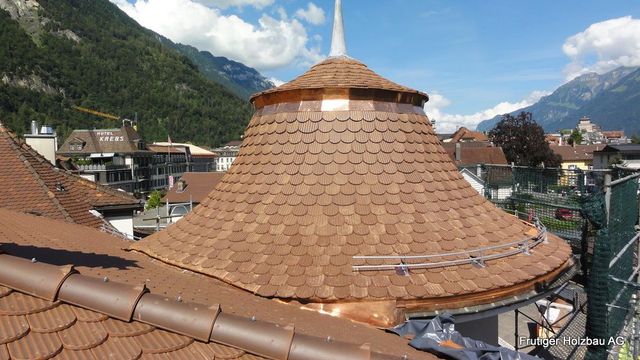 Weisses Gebäude mit rotem Ziegeldach und Dachgauben, blauen Fensterläden und Schornstein. Bewölkter Himmel, im Hintergrund ein Berg.