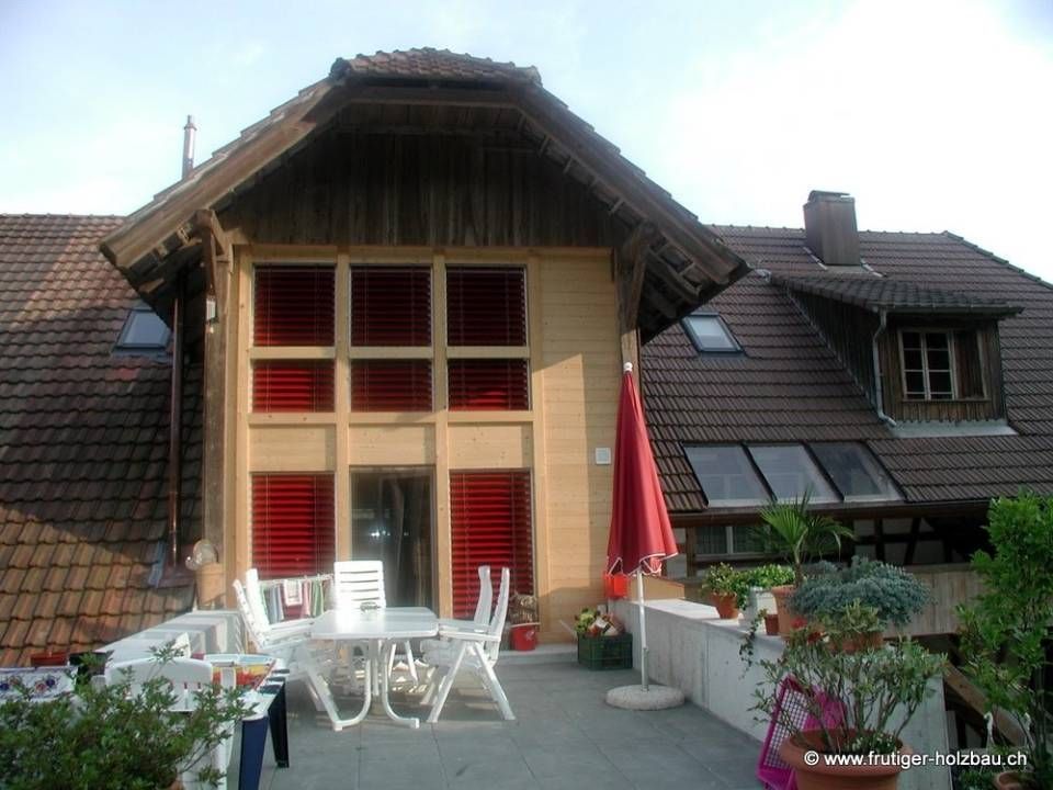 Terrasse mit weissen Möbeln vor einem Gebäude mit roten Fensterläden und braunem Dach.