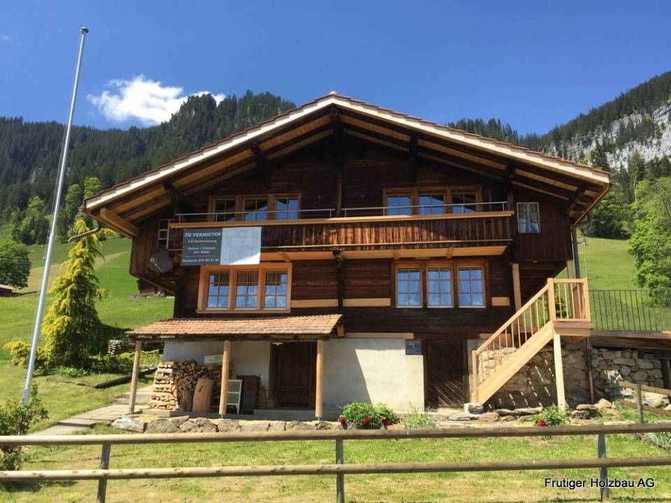 Holzhaus im Chalet-Stil mit Balkon und Treppe, eingebettet in eine grüne Hügellandschaft mit Bergkulisse.